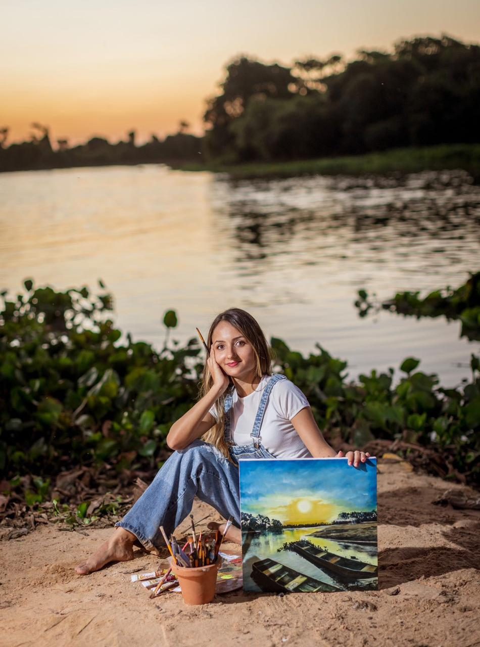 Adaiele Almeida sentada na areia à beira do rio, ao pôr do sol, ao lado de uma tela com barcos e o sol se pondo.